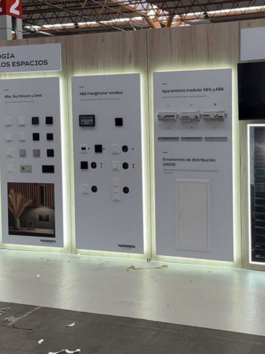 Stand de exposición de la marca Niessen en una feria comercial, con paneles retroiluminados que muestran mecanismos eléctricos decorativos, sistemas domóticos inalámbricos y cajas de distribución modular.