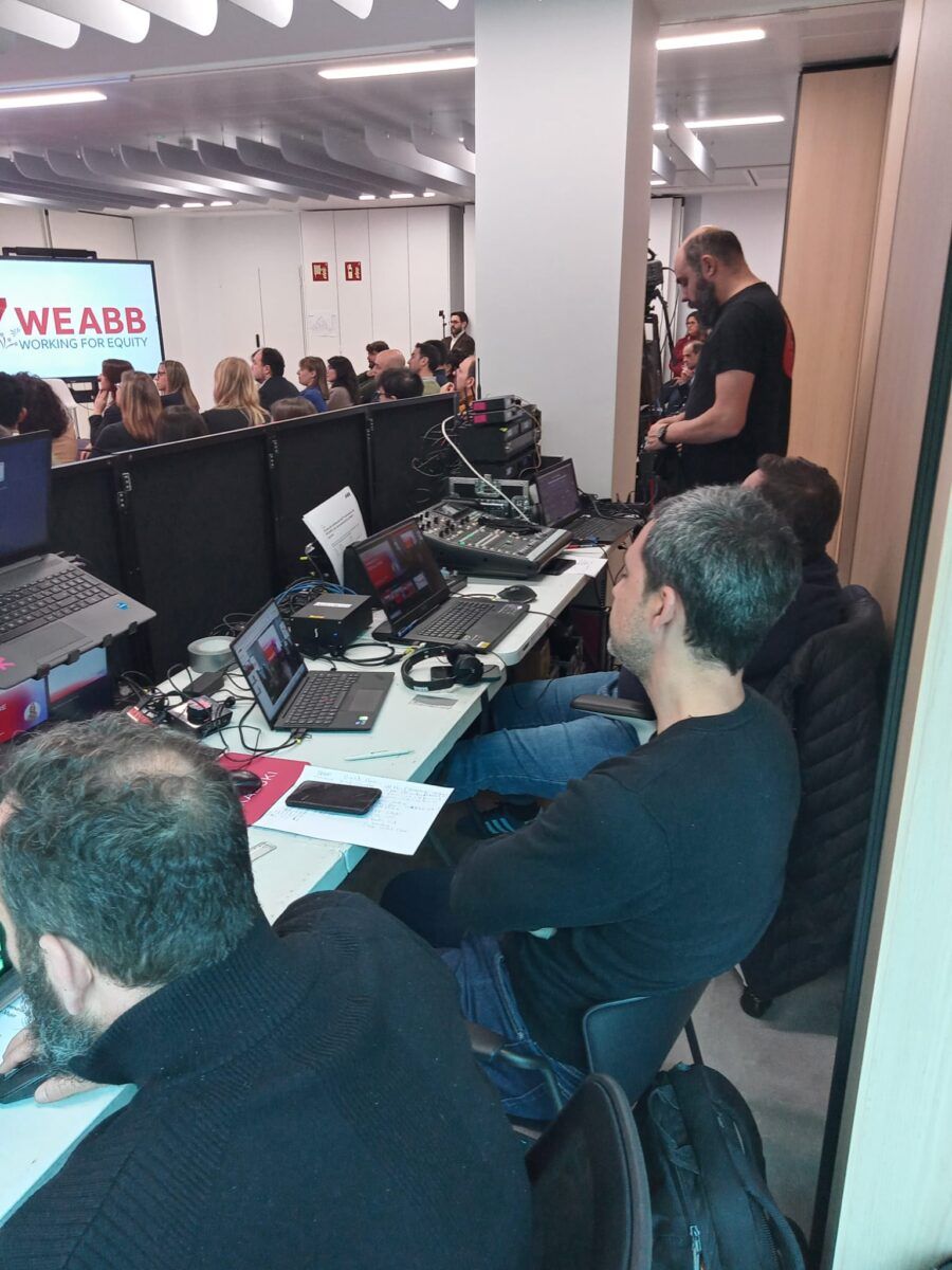 Vista de la cabina técnica durante un evento de la empresa ABB. Varios operadores de vídeo y sonido supervisan la transmisión en ordenadores portátiles, mesas de mezclas y pantallas de control. Al fondo, se ve al público asistente y una pantalla con el logo de WEABB.