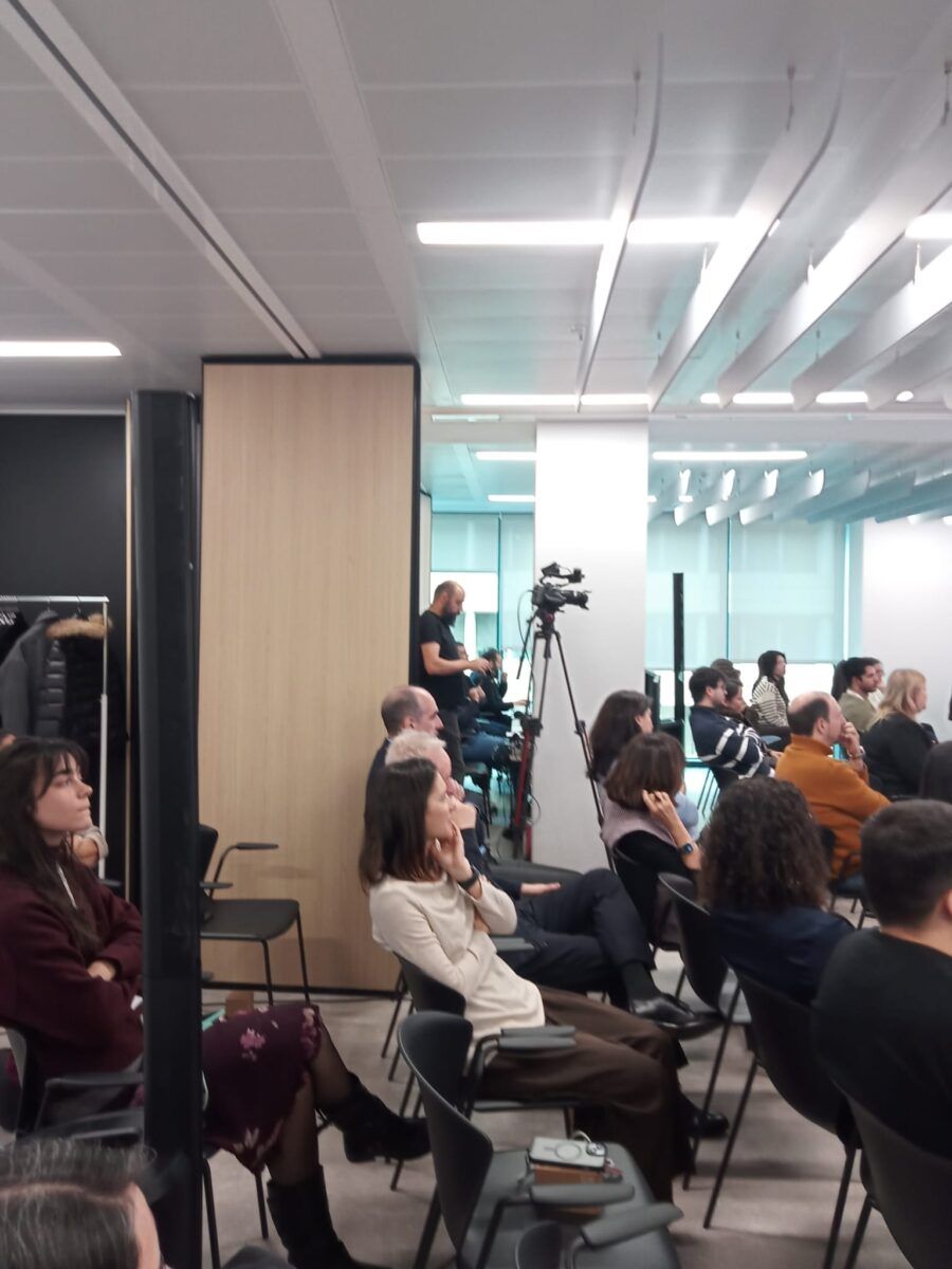 Asistentes sentados en un auditorio corporativo moderno de oficinas escuchando una ponencia. Entre el público se observa una cámara de vídeo sobre un trípode alto para la grabación y retransmisión en directo del evento.